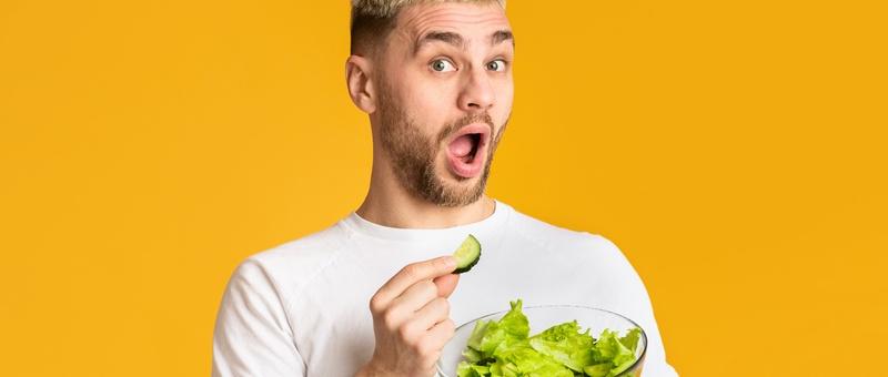 Man in white t-shirt eating a salad bowl with his hands against a yellow background.