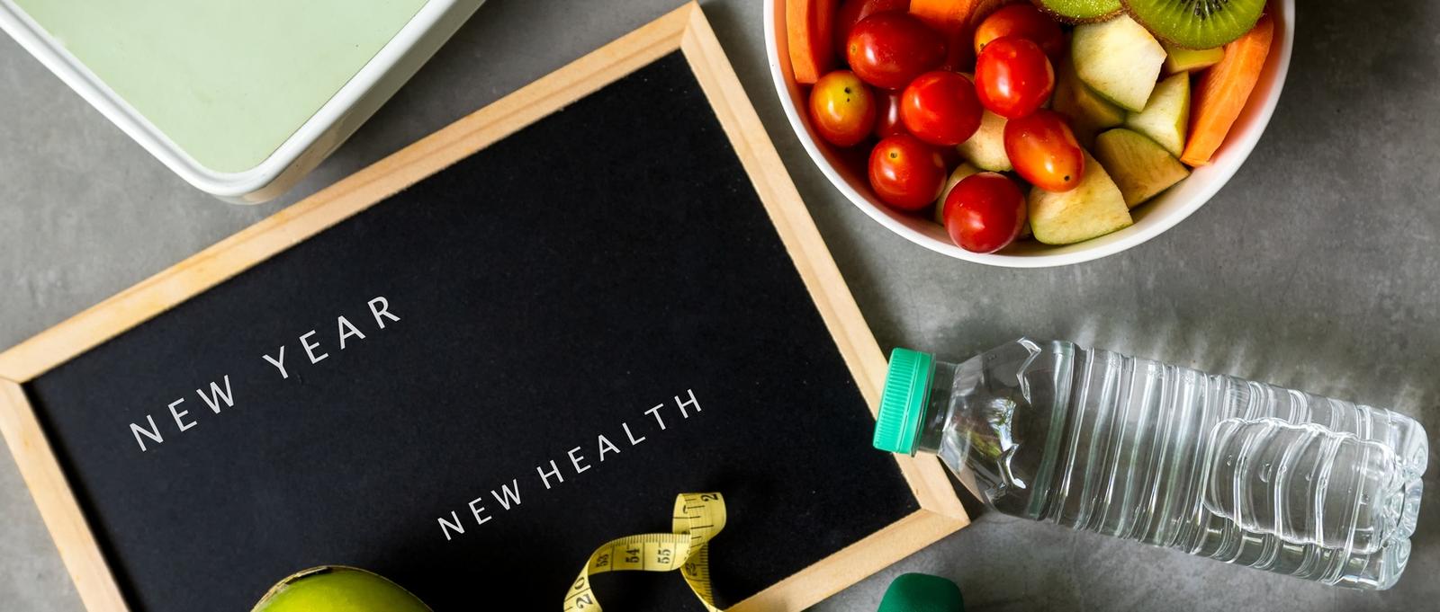 Blackboard with "NEW YEAR NEW HEALTH" text, fresh fruit bowl, water bottle, and measuring tape for healthy resolutions.