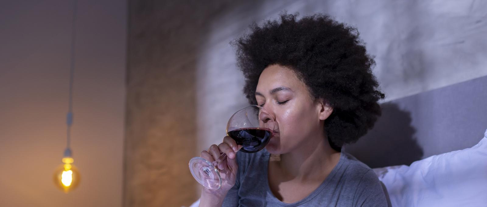 Person with curly hair sipping red wine from a glass in a softly lit room with a warm pendant light visible.