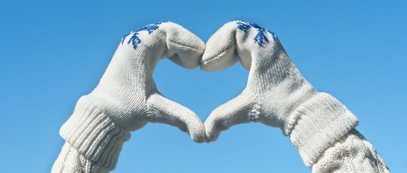 Person making heart hands wearing knitted white jumper, and knitted white gloves with snowflake prints, against a wintery blue sky.
