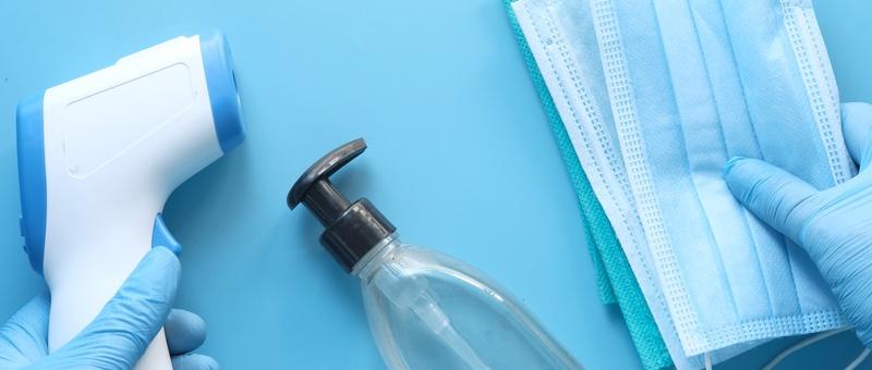 Surgically gloved hand holding blue medical face mask, infrared thermometer, and bottle of sanitising gel against blue background.