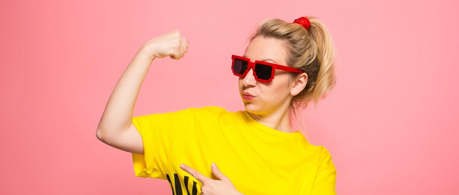 Woman in yellow T-shirt, red jeans and sunglasses showing her muscles against a pink background.