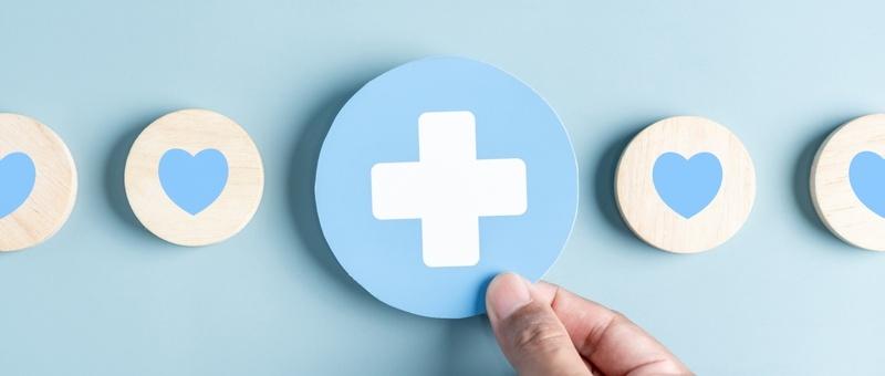 Hand holding white and blue pharmacy cross between blue and white hearts against a blue background.
