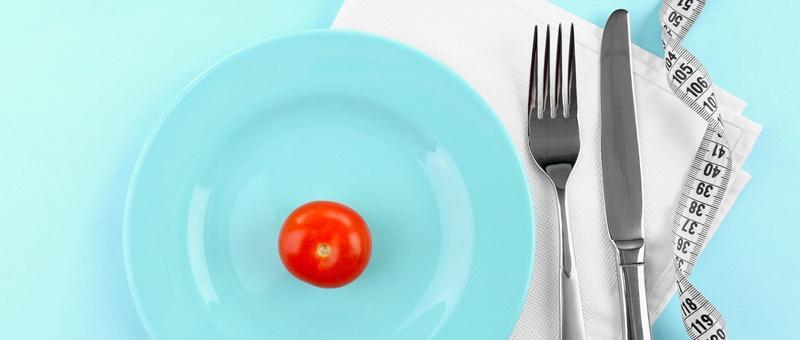 Red cherry tomato on a white plate beside measuring tape and cutlery. Blue background.