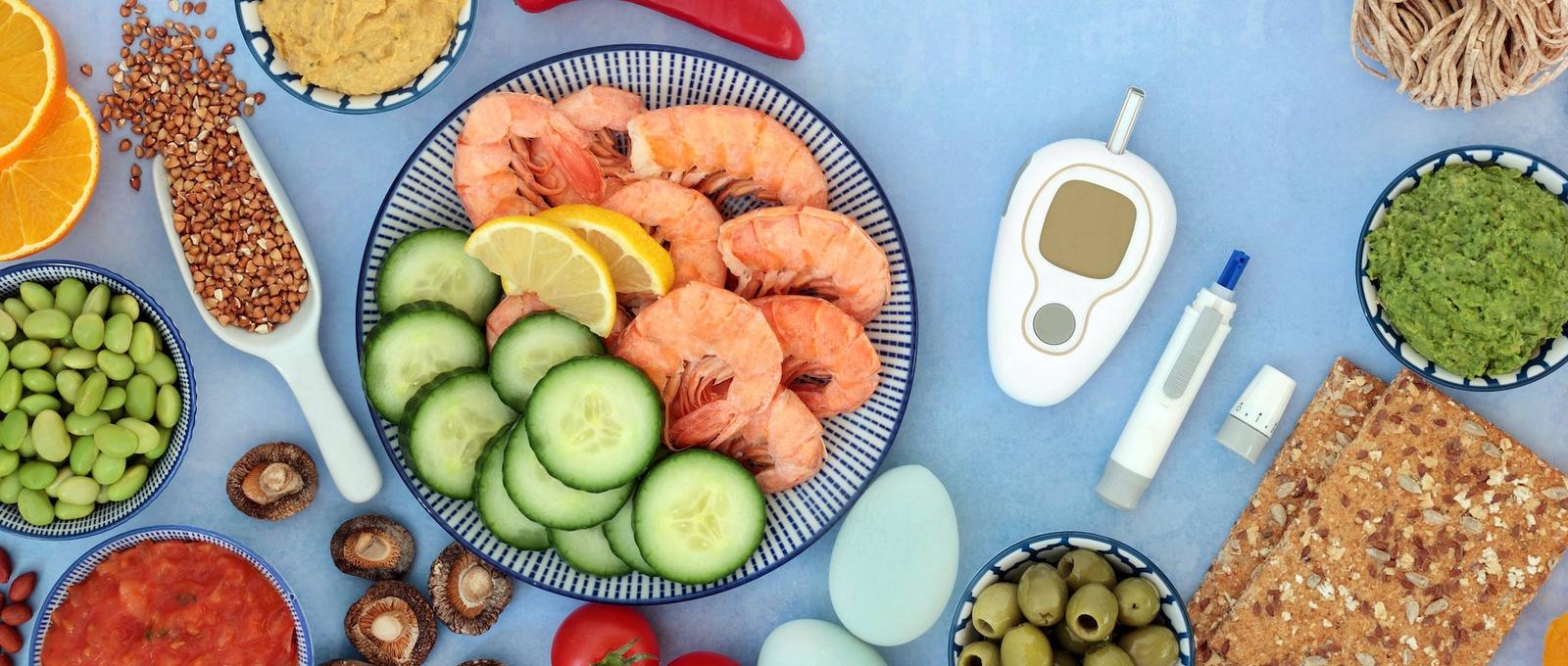Healthy diabetic meal with prawns, cucumber, and various foods alongside a glucose meter and insulin pen on blue background.