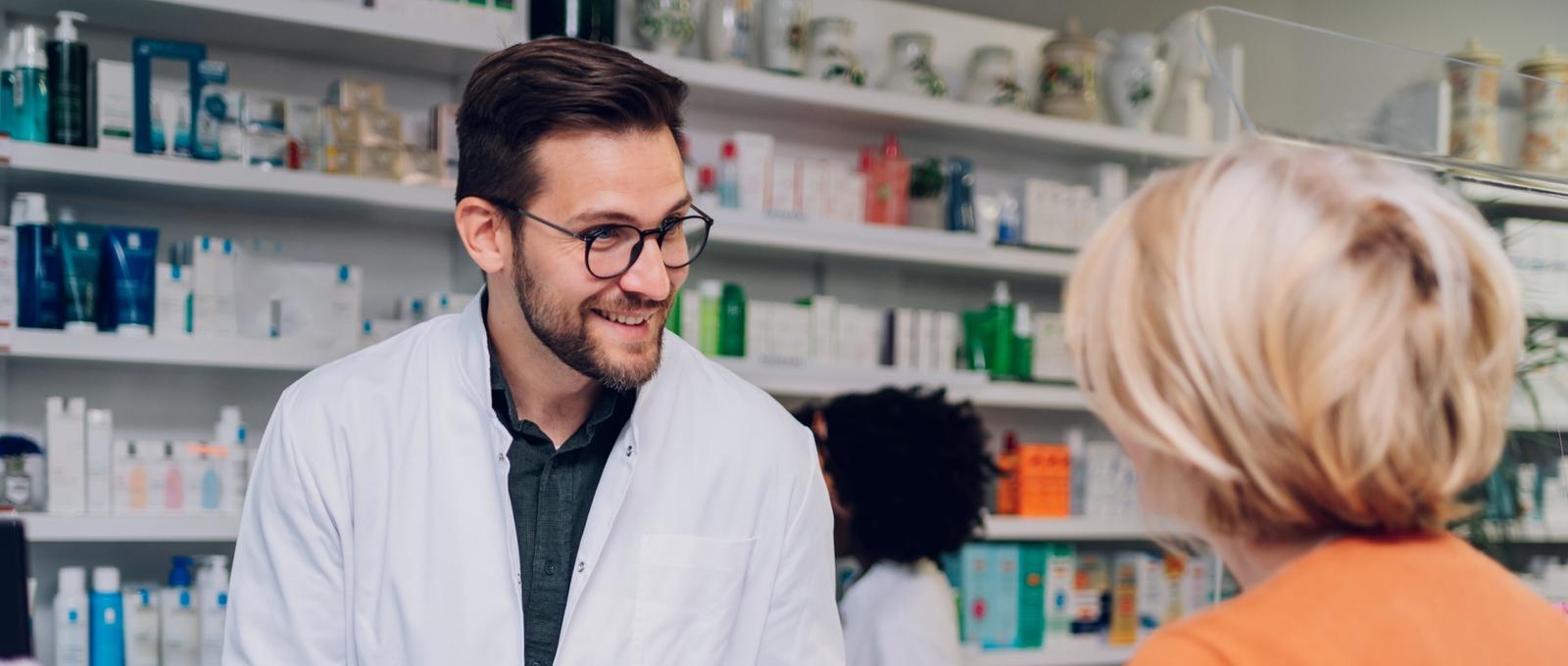 Young male pharmacist talking to female customer in a pharmacy with female pharmacist in the background. Shelves with health care products.