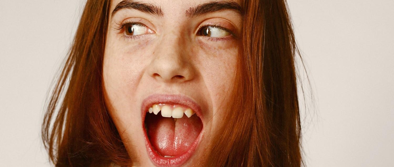 Close-up portrait with auburn hair showing an open mouth and eyes looking sideways against a neutral background.