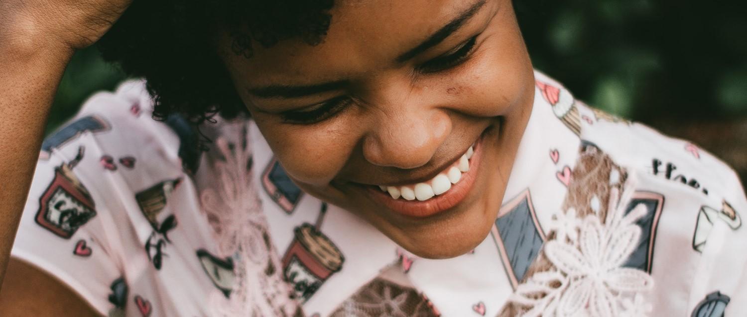 Close-up of a person with curly hair smiling warmly, wearing a patterned white shirt with illustrated designs.