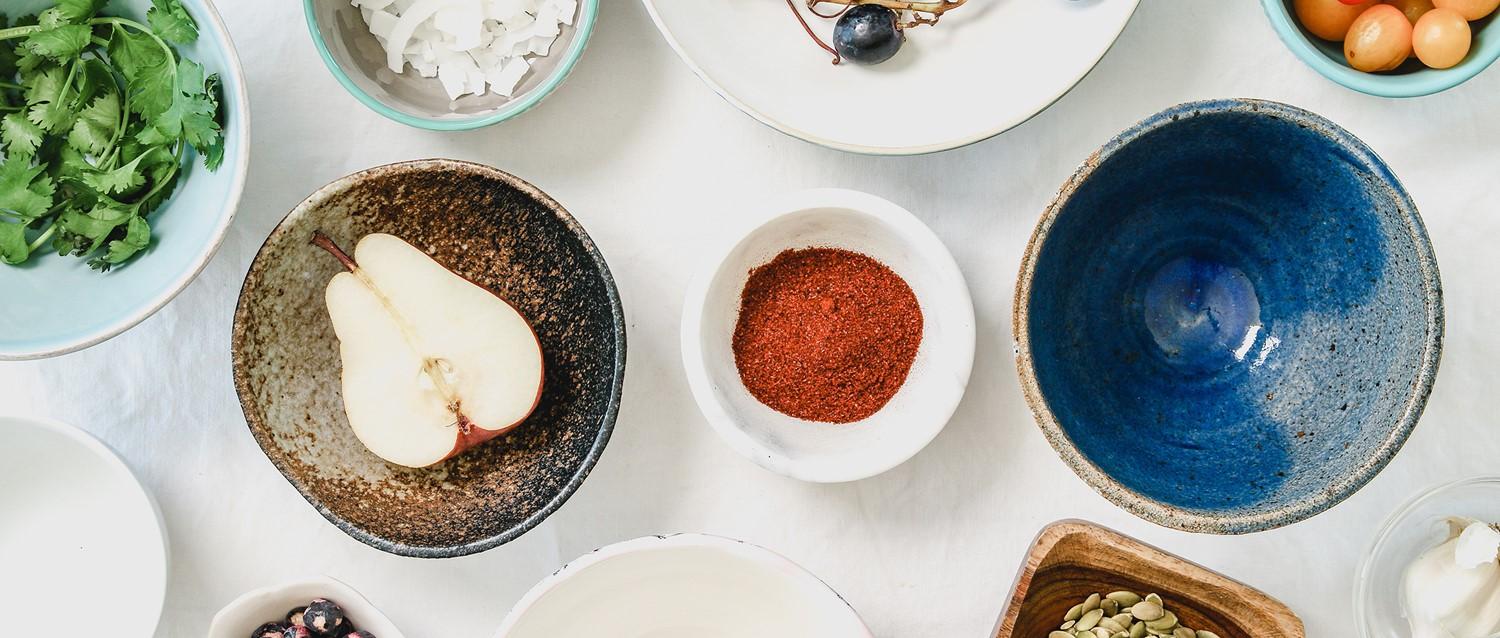 Overhead view of ceramic bowls containing fresh ingredients including a pear, red spice, herbs and a blue glazed bowl.