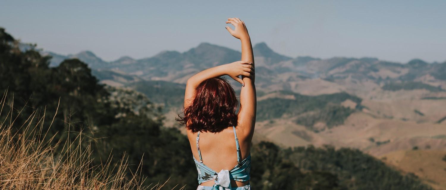 Person with auburn hair raising arm towards mountain landscape, viewed from behind in summer attire.