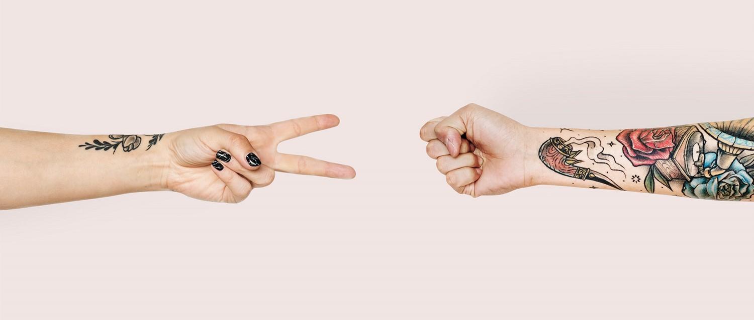 Two tattooed arms facing each other - one making scissors gesture with black nails, the other a closed fist with colourful sleeve.