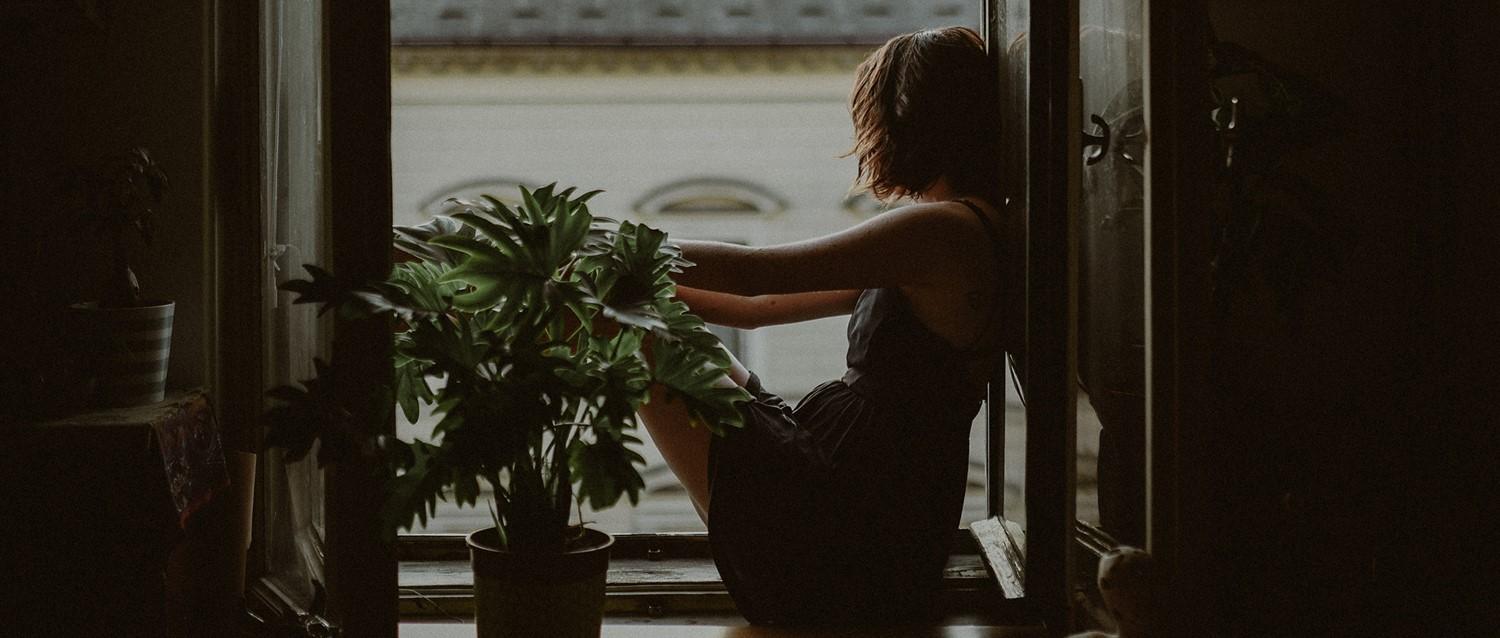 Person sitting in dim window light, tending to houseplants, creating a peaceful, contemplative atmosphere.
