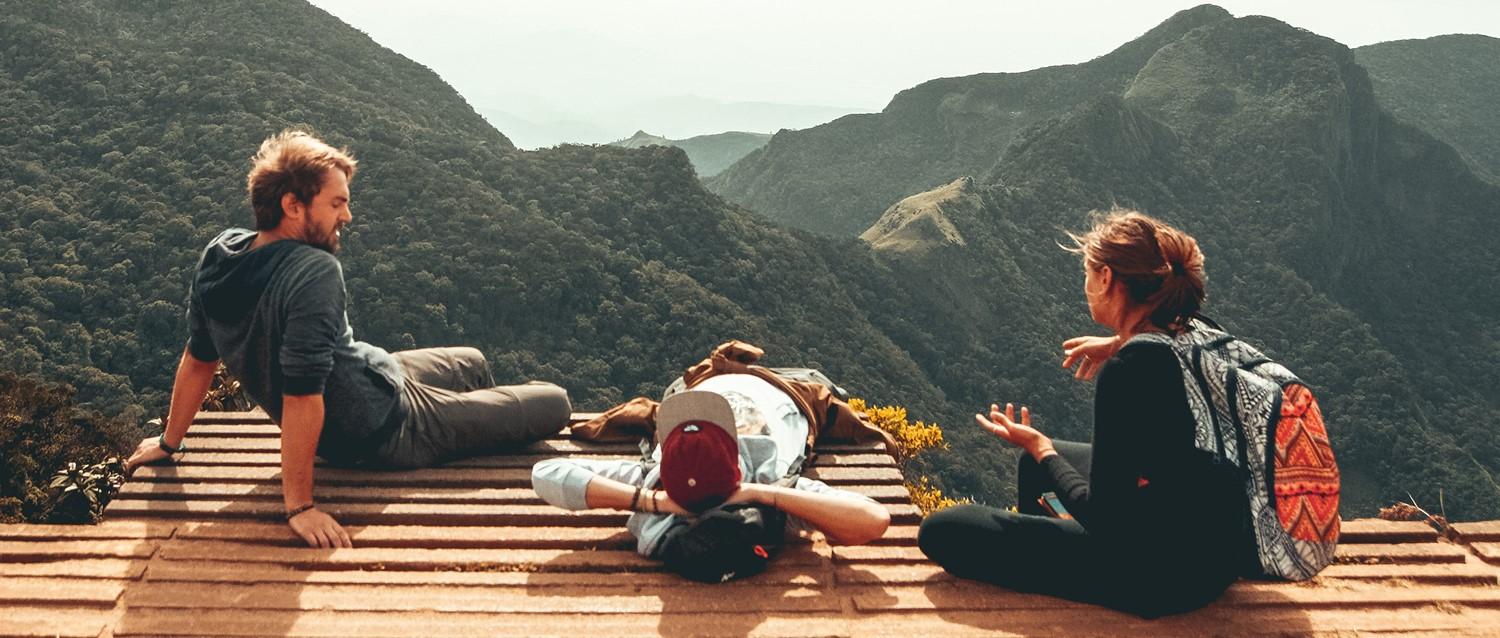 Hikers resting on wooden platform overlooking dramatic mountain landscape with lush green valleys and distant peaks.