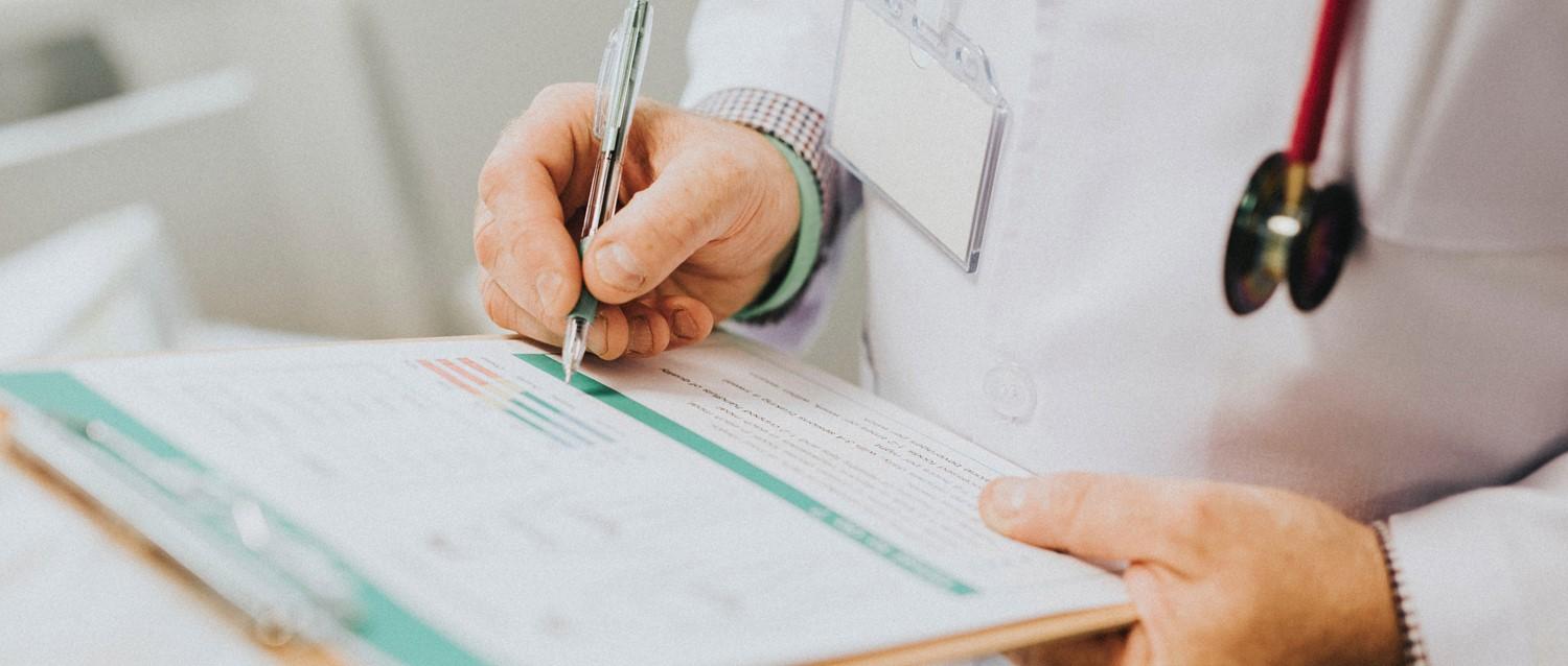 Doctor with stethoscope writing on medical chart, wearing white coat with ID badge and checkered shirt sleeve.