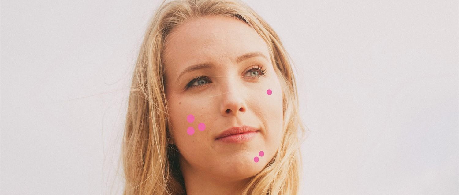 Person with blonde hair against light background, with small pink dots on their face in a pattern.