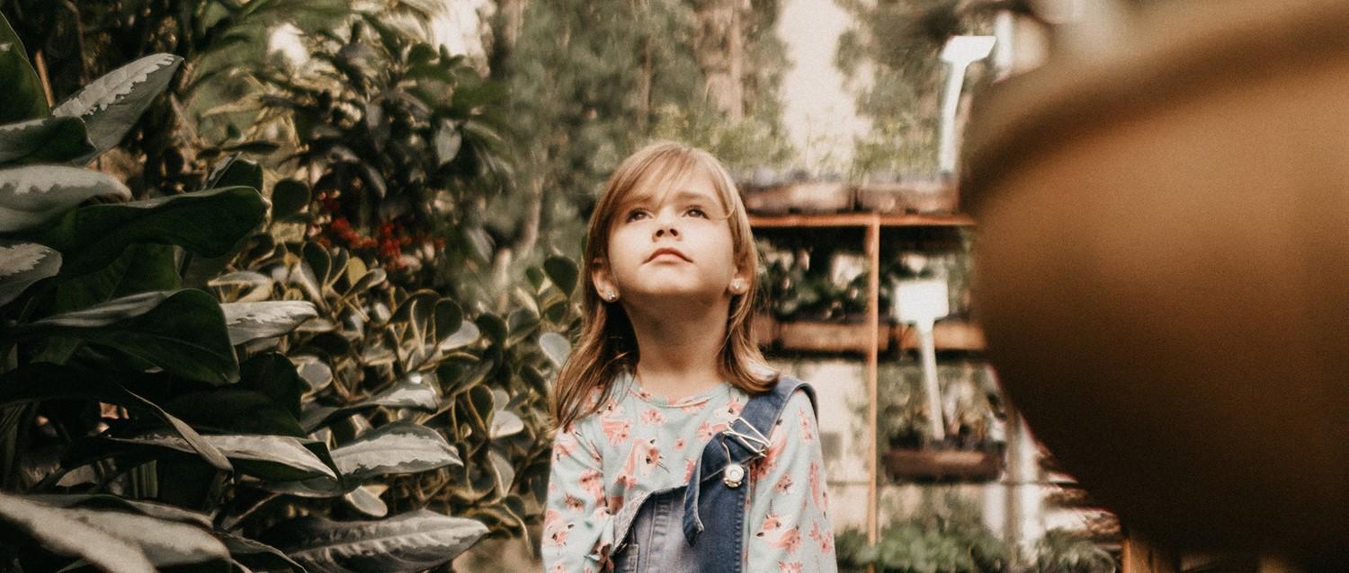 Young child in floral top and denim overalls looking upward in a lush garden setting with green plants surrounding.