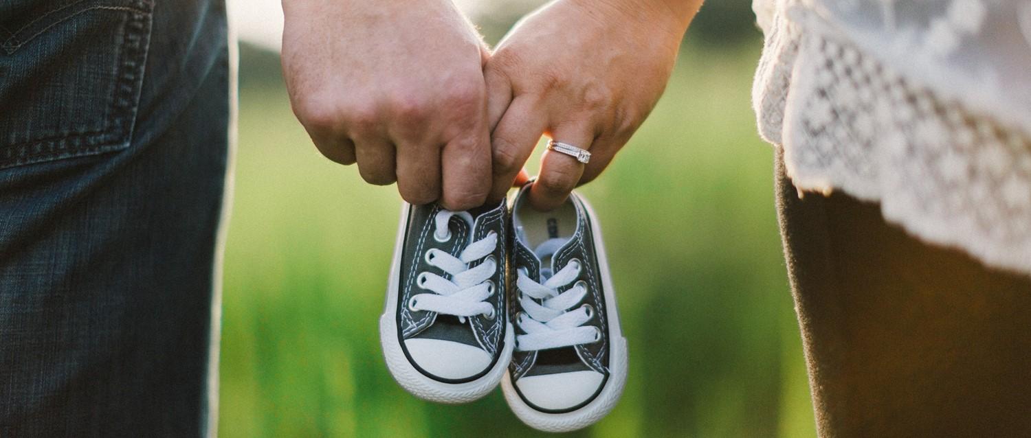 Couple holding hands and tiny black baby shoes, with one person wearing an engagement ring against a green background.