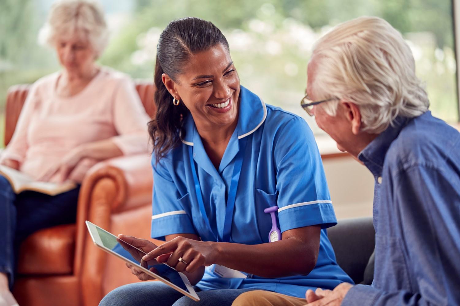 Healthcare worker in blue uniform smiling while showing a tablet to an elderly person in a care home setting.